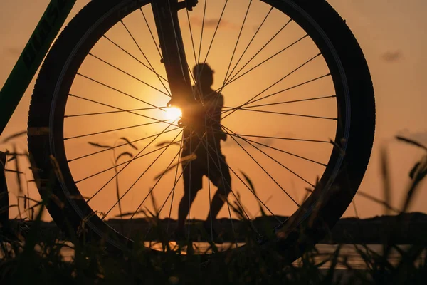 Close-up silhouette of a bike wheel at sunset. The sun shines through the wheel of a bicycle with blurred  runner figure at background