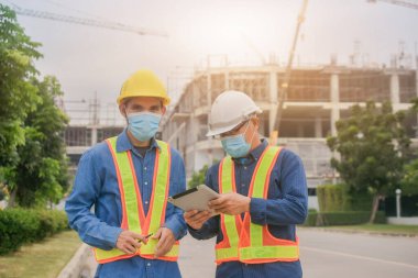 Yüz maskesi takan iki mühendis, inşaat maskesinde çalışan tablet kullanıyor. Coronavirus Covid19 'u koruyor.
