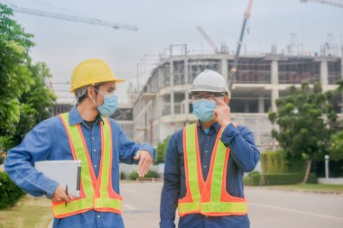 Yüz maskesi takan iki mühendis, inşaat maskesinde çalışan tablet kullanıyor. Coronavirus Covid19 'u koruyor.