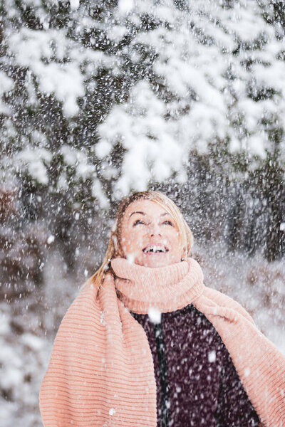 Blonde woman with purple coat and pink scarf enjoys of heavy snowing and blizzard during winter time in forest background