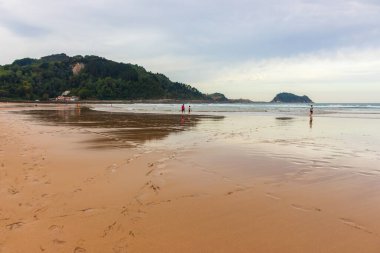 10 / 12 / 2019 - İspanya: Biscayne Körfezi 'nin geniş sahillerinde yürüyen insanlar. Sörfçülerin olduğu sörf sahili ve arka planda dağ. Aktif yaşam tarzı konsepti. Sakin resimli deniz kıyısında. Deniz kıyısında bulutlu bir gün