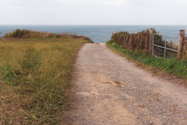 Denize giden boş bir yol. Camino de Santiago konsepti. Kırsal alan. Çayır boyunca sahile giden patika. Yürüyüş ve seyahat konsepti. Kırsal doğa. Bakış açısına göre köy yolu. Arka planda okyanus olan sakin bir manzara.