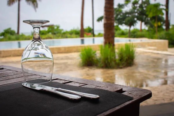 Dinner table with glass and knife. Empty outdoor restaurant near pool. Wineglass on wooden table in resort cafe. Tropical resort background. Food and drink concept. Empty resort.