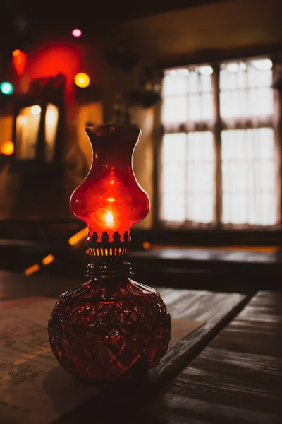 Small oil lamp on the table. Old oil lantern with window on background. Red kerosene lamp. Cozy rustic interior. Evening concept. Ancient decorative lamp on table. Vintage home objects. Home decoration.