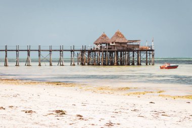Güneşli bir günde teknesi ve sahil kulübesi olan ahşap iskele. Hint Okyanusu 'nun alçak gelgiti. Idyllic egzotik tatil köyü. İskele, bungalovlara bakıyor. Tropik cennet. Göl kenarında balayı. Sahne sakin deniz manzarası. Adadaki sahil kulübesi. Yaz seyahati.