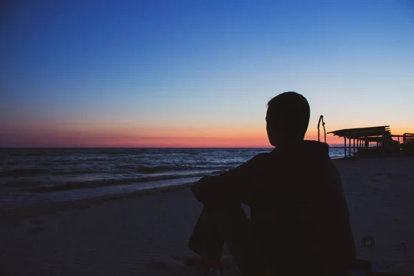 Silhouette of unknown person watching the sunset over the sea. Unknown tourist on coast against idyllic evening sky. Beautiful sunset over sea with sitting man on the beach. Summer vacations. Bright evening sky with man silhouette.