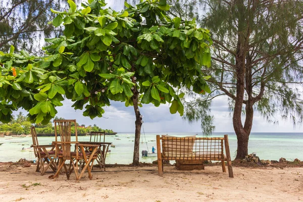 Wooden chairs and table under trees on tropical beach. Outdorr cafe furniture in resort garden against seascape. Bamboo bench and chairs on beach. Idyllic leisure on seacoast. Vacations in Africa. Sunny day in paradise.