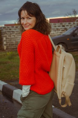 Sırt çantalı parlak turuncu kazaklı esmer kadın. Modern sonbahar modası. Örgü elbiseli şirin bir kız. Büyük şehir hayatı. Sokak hayatı. Vahşi ve özgür bir kadın. Sıcak giysiler içinde gülümseyen kadın..