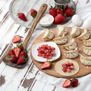 Taze çilekler yuvarlanan hamur çemberleriyle doluyor. Vejetaryen yemeği pişirme süreci. Hamur köftesi hazırlamak için gerekli malzemeler ve mutfak aletleri. Üst Manzara. 1x1 biçimi