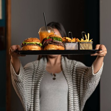 Fast food menüsündeki tepsiyi tutan kadın. Büyük hamburgerler, patatesler ve plastik bardakta taze portakal suyu. Besleyici atıştırmalıklar. Misafirperver bayan klasik Amerikan yemeği yer. Lezzetli ama sağlıklı değil.