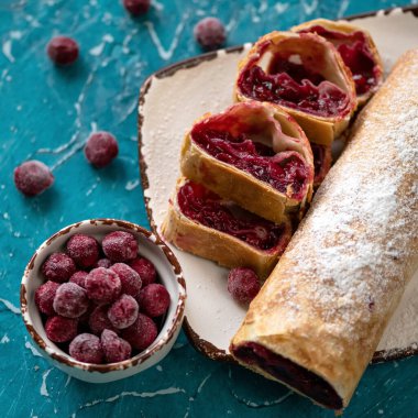 Kirazlı çörek, turta ya da turta. Gurme tatlı, dondurulmuş böğürtlenlerle doldurulmuş tatlı hamur işleri mutfak masasının mavi arka planında. Tabağa toz şeker serpiştirilmiş dilimler ve rulo. Üst görünüm