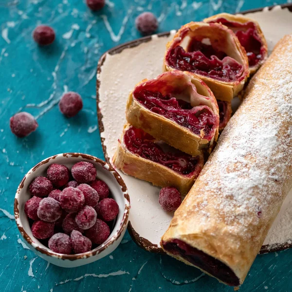 Kirazlı çörek, turta ya da turta. Gurme tatlı, dondurulmuş böğürtlenlerle doldurulmuş tatlı hamur işleri mutfak masasının mavi arka planında. Tabağa toz şeker serpiştirilmiş dilimler ve rulo. Üst görünüm