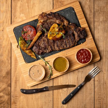 Izgara sebzeli büyük et bifteği ve ahşap kesim tahtasında soslar ya da çatal ve kaşık dolu bir masa. Yürekli et yemeği yemeye hazır. Üst manzara, yumuşak odaklanma. Pankart için kare biçimi