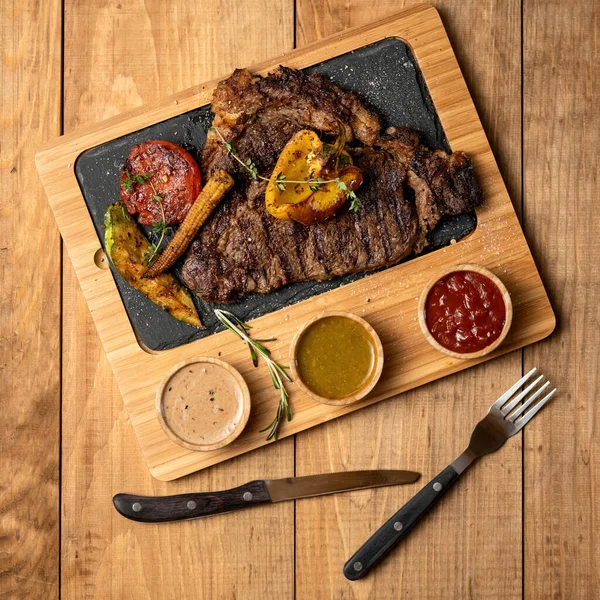 Izgara sebzeli büyük et bifteği ve ahşap kesim tahtasında soslar ya da çatal ve kaşık dolu bir masa. Yürekli et yemeği yemeye hazır. Üst manzara, yumuşak odaklanma. Pankart için kare biçimi