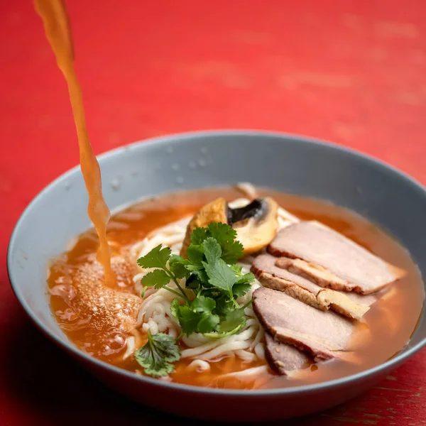 A sopa de ramen vaza-se com o caldo. Tigela de macarrão, carne e salsa de ervas na mesa vermelha. Prato oriental picante, cozinha pan-asiática — Fotografia de Stock