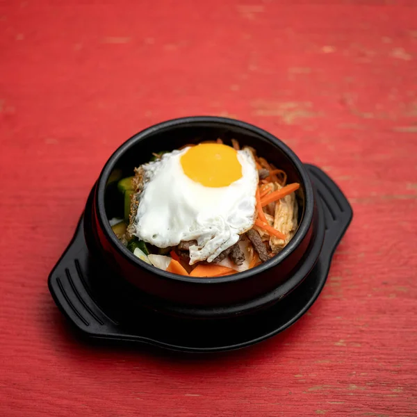 Pan-Asian cuisine. Black clay pot with food on red background. Dish of vegetables and meat, garnished with fried egg on top. Spicy oriental dish — Stock Photo, Image