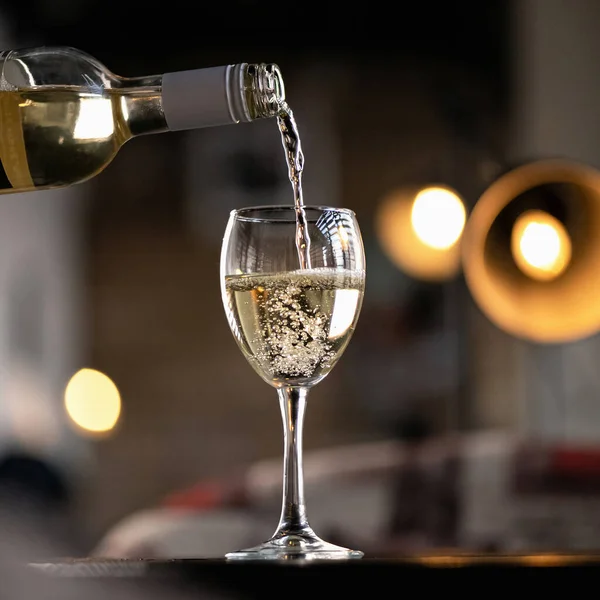 Белое вино наливается из бутылки в бокал вина. Алкогольный напиток с пузырьками воздуха на столе на подсветке размытого фона. Вид сбоку — стоковое фото