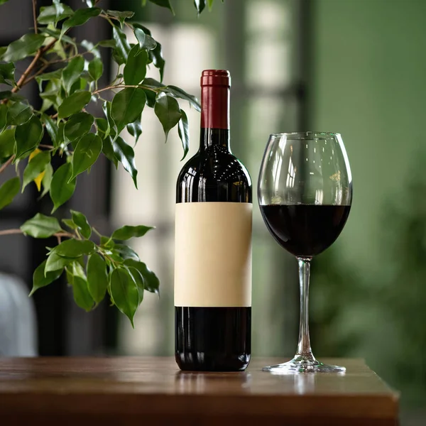Придумайте или искушайте дизайн этикетки вина. Бутылка и бокал с красным вином за деревянным столом против интерьера комнаты на размытом фоне с зеленым фикусом слева. Алкогольные напитки — стоковое фото