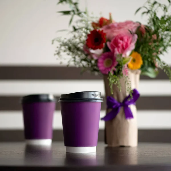 Coffee drink in paper cups on table. Purple disposable cardboard cups and bouquet of roses on blurred background. Coffee to go concept. Soft focus. Square format with copy space. Blank paper cup