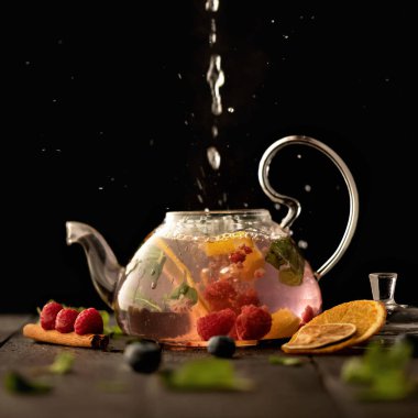 Cam çaydanlığa akan su akıntısı ve siyah arka planda böğürtlen çayı. Tarçın çubuğu, ahududu ve yaban mersininden, nane yapraklarından ve bir dilim portakaldan iç. Çay töreni konsepti