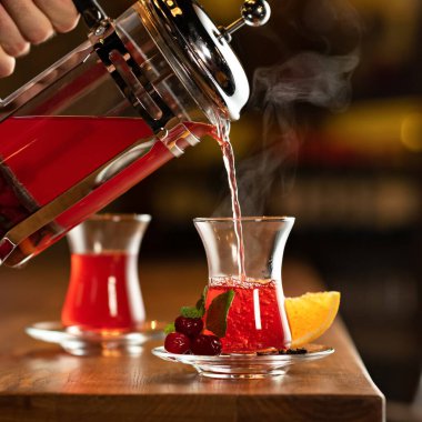 Çaydanlıktan fincana sıcak kiraz çayı dökülüyor. Çay töreni konsepti. Erkek el, şeffaf Fransız presinden bardağa su döker. Yakın çekim. Yan görünüm