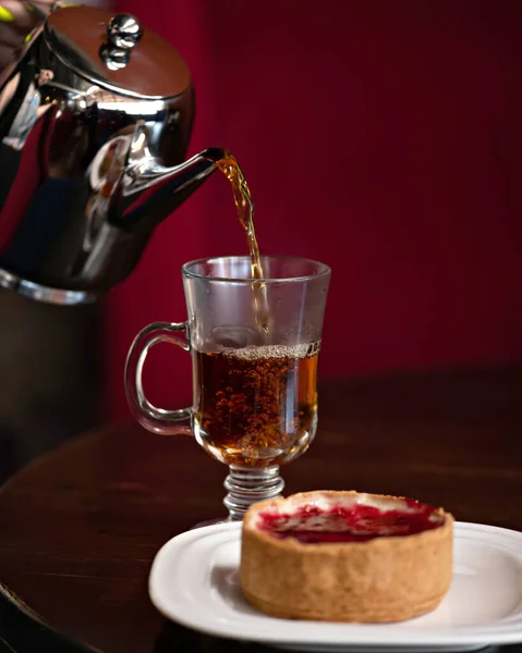 Heißer schwarzer Tee, der aus einem Wasserkocher in eine Glasschale auf dem Tisch mit Kirschdessert gegossen wird. Konzept der Teezeremonie. Kopierraum — Stockfoto