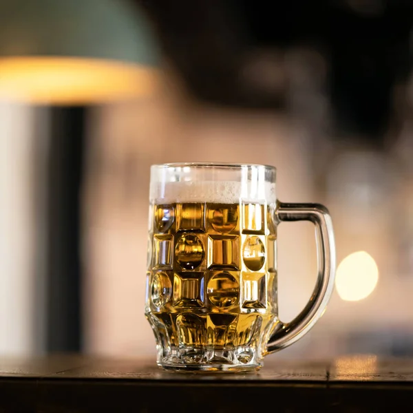 Glass cup of chilled light beer with froth. Alcoholic beverage on counters of brewery against of backlit blurred background. Side view. Copy space for text — Stock Photo, Image