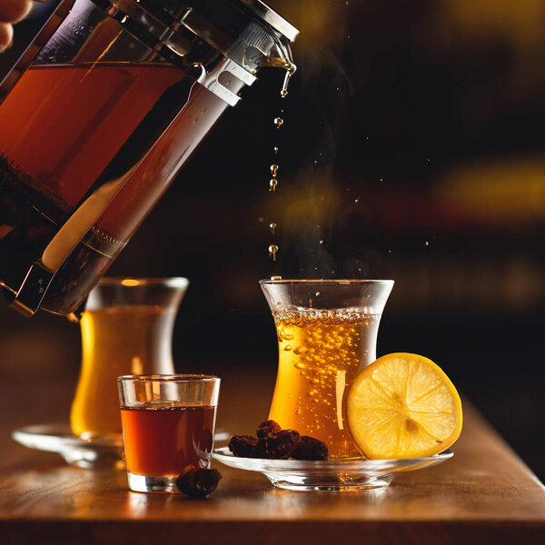 Налить черный чай Баклит. Золотой напиток течет из прозрачного чайника в стекло на размытом фоне. Вид сбоку. Концепция чайной церемонии. Скопируйте пространство справа. Формат квадрат. Мягкий фокус