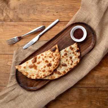 İki lezzetli fırınlanmış cheburek, etli gevrek puf böreği. Düz yatıyordu. Ahşap masa örtüsüne doldurulmuş hamurdan yapılmış tabakla çatal bıçak takımı ve tepsi. Kare biçimi