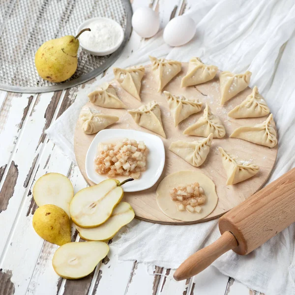 Cooking process. Sweet Dumplings with pear filling on Raw dough circles at wooden cutting board. Convenience Food concept. Ingredients and rolling pin on rustic background. Square format