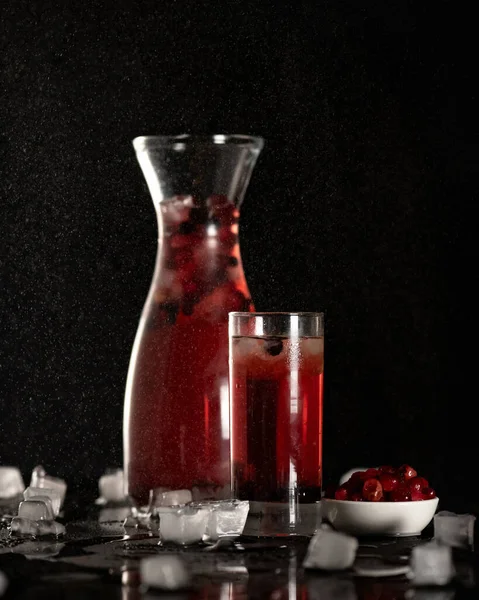 Склянка та глечик вишневого напою на столі з замороженими ягодами та кубиками льоду. Мокрий скляний посуд з червоним безалкогольним напоєм на чорному тлі з розбризкуваними краплями води. Вид збоку. М'який фокус. Копіювати простір — стокове фото