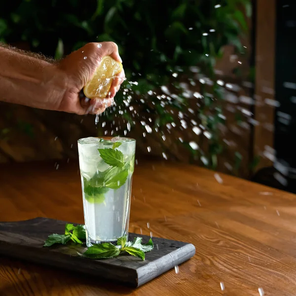 Juice splashes or drops in motion. Man squeezing lemon juice into glass with mint lemonade on wooden table. Healthy refreshing drink, Cooking or preparing process. Soft focus. Copy space — Stock Photo, Image