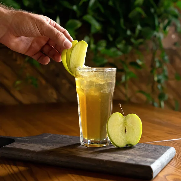 Шеф-повар украшает яблочный сок или пьет стакан зелеными ломтиками яблока. Мужская рука в процессе приготовления фруктового освежающего напитка. Витамины от природы на деревянном столе, размытый фон. Мягкий фокус — стоковое фото