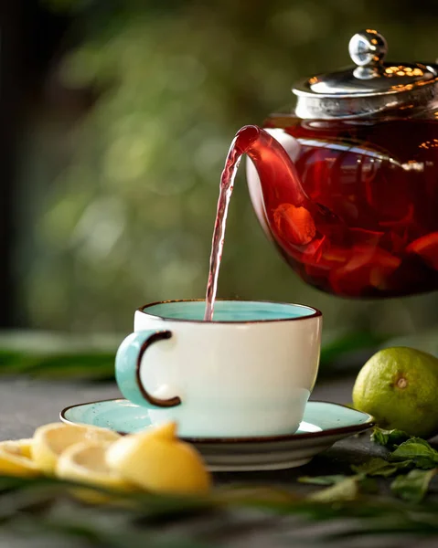 Schwarzer Tee mit Zitrone und Minze gießt aus einer glasklaren Teekanne in eine Tasse oder einen Becher auf dem Tisch. Objekte auf verschwommenem grünem Hintergrund. Seitenansicht — Stockfoto