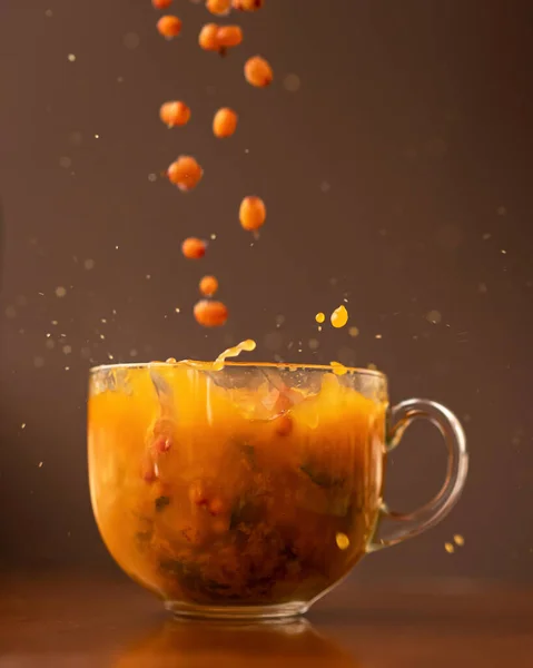 Морський пряний чай. Ягоди падають у скляну чашку з помаранчевим трав'яним напоєм. Збільшений звук на коричневому фоні, м'який фокус. Сторона зору — стокове фото