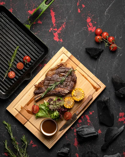 Hot Meat t-bone steak, ribeye or Tomahawk. Flat lay. Cutting board with a hearty lunch and vegetables on a black table with grill pan and coals on it. Top view. Copy space