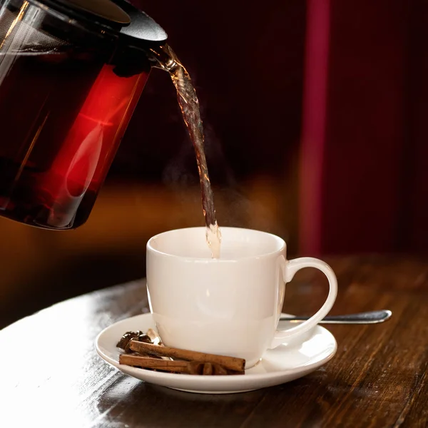 Morgenentspannung und eine Tasse Tee. Heißes gebrühtes Getränk, das in weiße Tasse auf Untertasse mit Zimtstangen darauf gegossen wird. Unscharfer Hintergrund. Teezeremonie, Seitenansicht. Kopierraum — Stockfoto