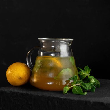 Nane ve limonata ferahlatıcı. Sarı içkili soğuk cam sürahi, siyah arka planda limon ve nane filizi. Yan görüş. Yumuşak odak