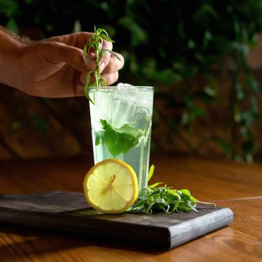 Adam ahşap masada naneli limonata yapıyor. Erkek eli, buzlu içeceği otlarla süslüyordu. Sağlıklı bir ferahlatıcı içecek, pişirme ya da hazırlama işlemi. Yumuşak odaklanma. Boşluğu kopyala