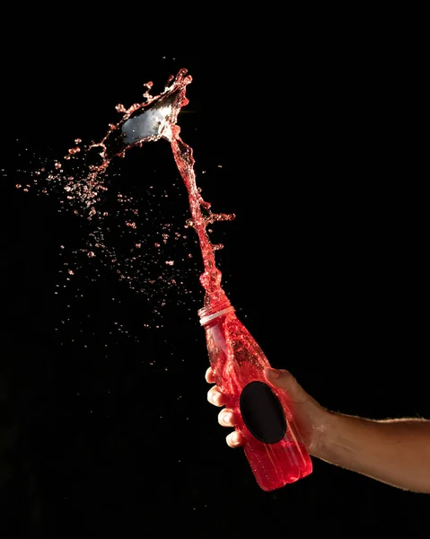 Rotes Getränk spritzt auf schwarzem Hintergrund. Hinterleuchtete männliche Hand, die eine durchsichtige Flasche mit schwarzem Blanko-Etikett hält. Seitenansicht. Leuchtende Farben. Mock-up oder Vorlage für die Etikettengestaltung. Kopierraum — Stockfoto