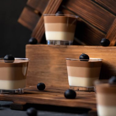 Üçlü çikolatalı mus tatlısı. Üç katmanlı siyah, süt ve beyaz çikolatalı, ahşap zemin üzerinde yabanmersini bardaklarıyla süslenmiş. Yumuşak odaklanma. Yakın çekim.