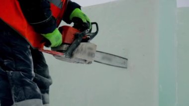 Koruyucu miğfer ve yansıtıcı yelek giyen bir işçi buz bloğunda benzopil ile bir delik açıyor.