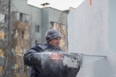 Mavi ceketli bir montaj işçisi ve siyah örülmüş bir şapka bir buz tabakasını testere ile düzeltiyor.