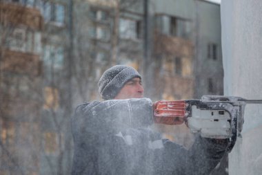 Mavi ceketli bir montaj işçisi ve siyah örülmüş bir şapka bir buz tabakasını testere ile düzeltiyor.