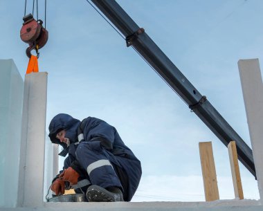 Mavi ceketli, kapüşonlu bir işçinin portresi. Buz kasabası inşaatında bir vinç kancasına takılmış.