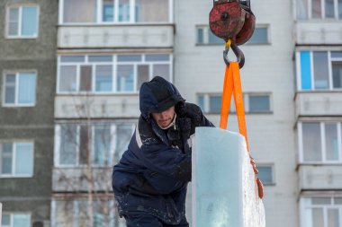 Mavi ceketli, kapüşonlu bir işçinin portresi. Buz kasabası inşaatında bir vinç kancasına takılmış.