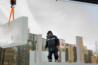 Mavi ceketli, kapüşonlu bir işçinin portresi. Buz kasabası inşaatında bir vinç kancasına takılmış.