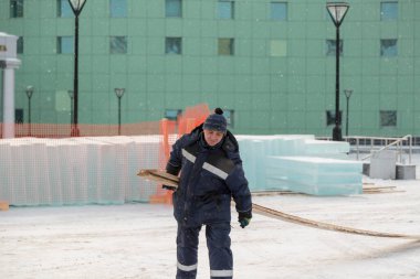 Elinde tahta olan mavi kapüşonlu bir müteahhit inşaat alanında yürüyor.