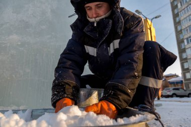 Kış tulumu giymiş işçi bir derleyici, ıslak karı yalaktan metal kovaya koyar.
