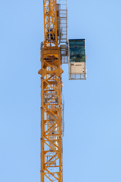 Fragment of yellow hoisting crane with cab on blue sky background.
